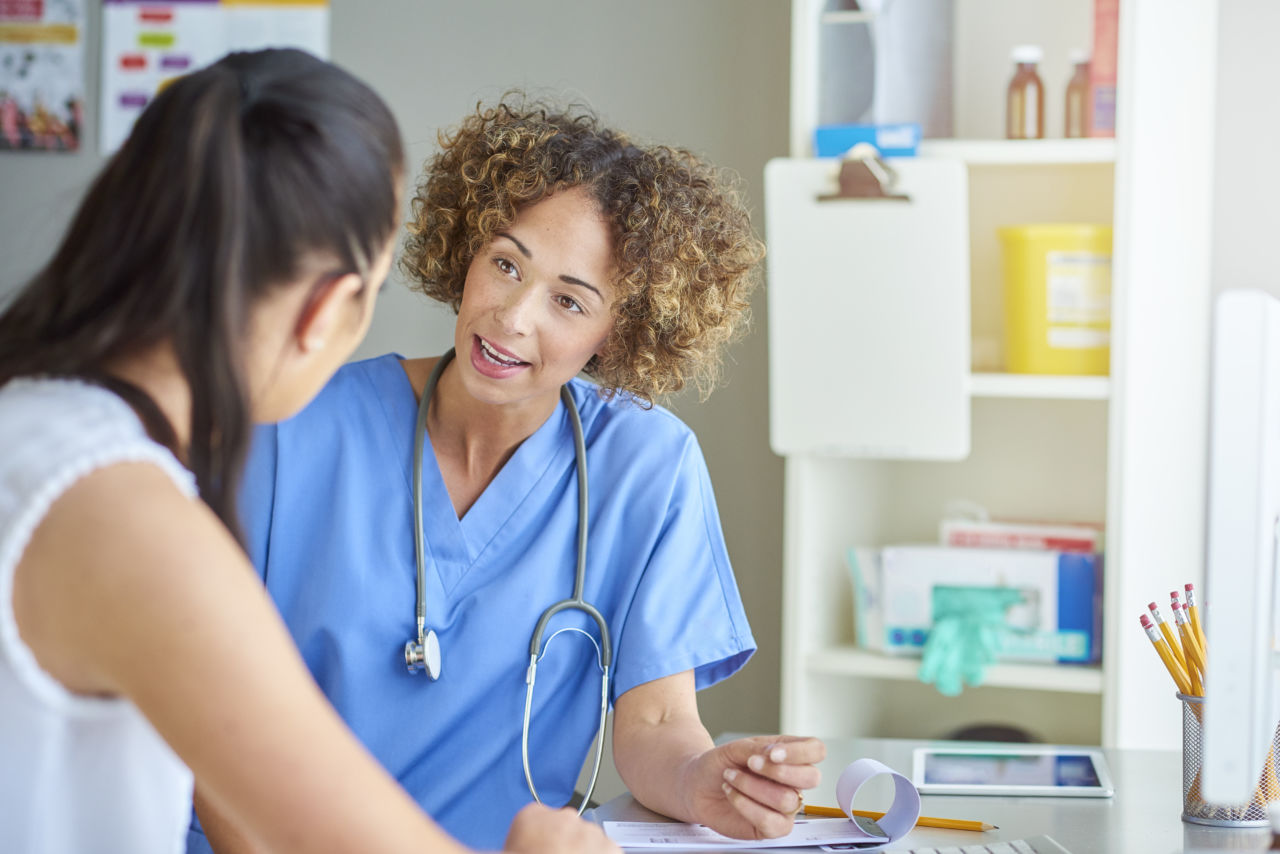 A female doctor sits at her desk and listens to a female patient .  She is wearing blue scrubs and stethoscope and is sitting at her desk . In the background shelving containing sharps box, gloves and medicine can be seen .